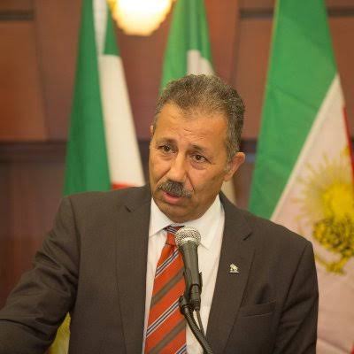Foad Pashaei, secretary-general of the Constitutionalist Party of Iran, speaking at a podium in front of Iranian flags.